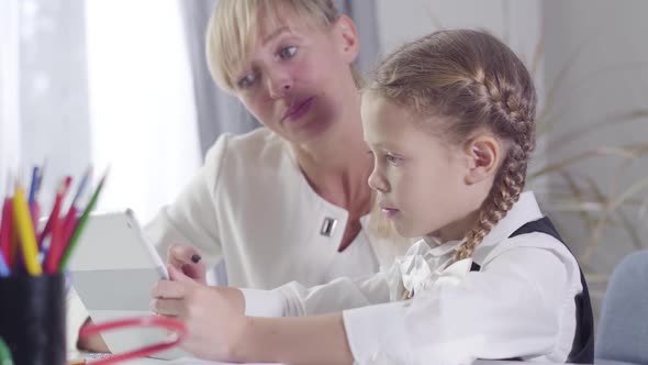 Side view of cute Caucasian schoolgirl sitting at the table with tablet as middle aged tutor explain