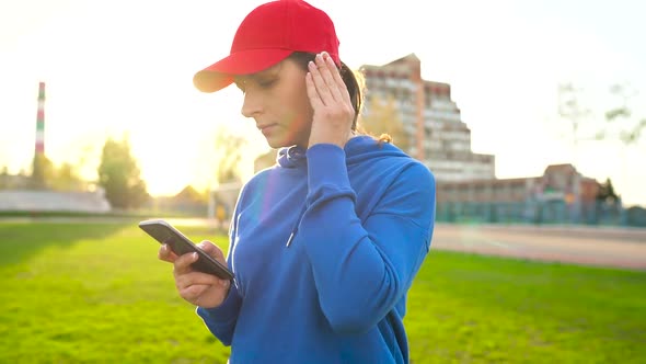 Woman with Wireless Headphones and Smartphone Chooses Music and Runs Through the Stadium at Sunset