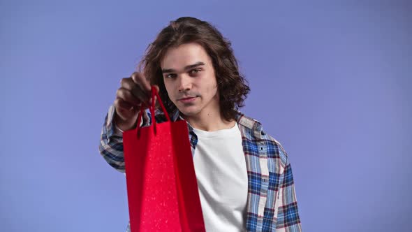 Charming Man Gives Red Gift Paper Bag By Hand to Camera on Violet Wall Background