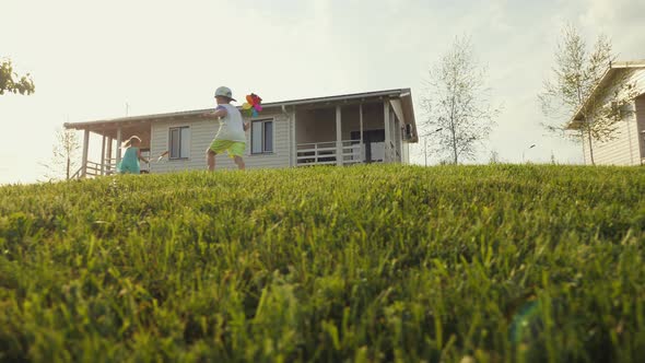 Happy Little Kids Running on Green Lawn and Holding Colored Windmill Toy