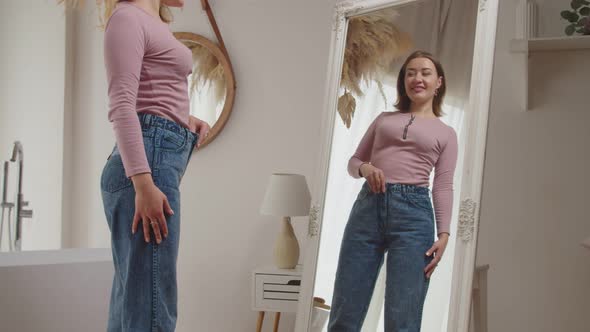 Reflection in Mirror of Pretty Adult Woman in Loose Pants Showing Her Weight Loss Success