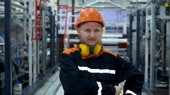 A Bearded Male Mechanic in an Orange Helmet Receives Instructions Over a Walkietalkie and Puts on