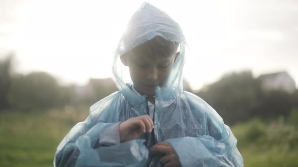 Cute Confident Boy Zipping Raincoat and Looking at Camera Standing on Rainy Summer Day Outdoors