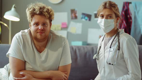 Portrait of Patient and Doctor in Mask Sitting on Sofa at Home