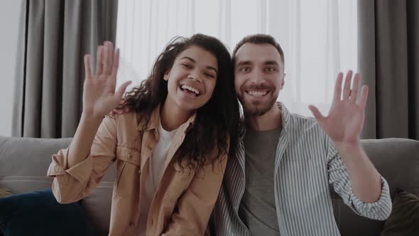 Portrait of Happy Multicultural Couple Sitting on Couch at Home Waving Hands at Camera Friendly