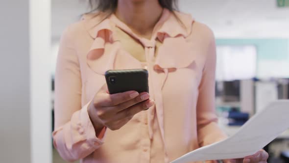 Video of midsection of biracial businesswoman using smartphone and walking in office