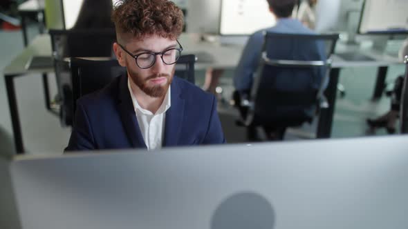 Serious Young Bearded Man Working on Decktop Computer While Working in Big Open Space Office