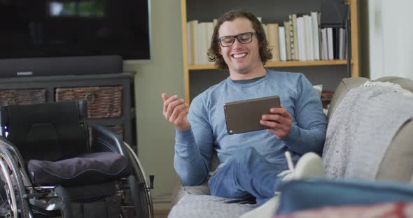 Happy caucasian disabled man sitting on sofa in living room using tablet