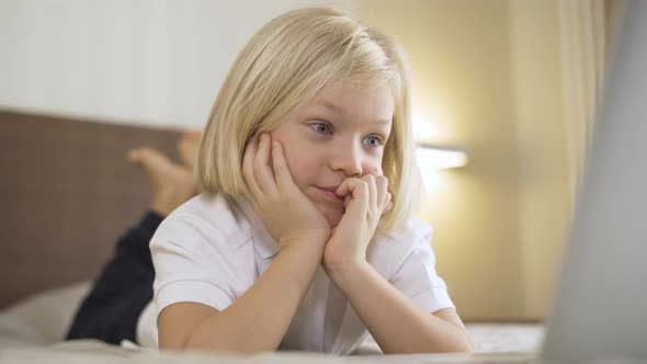 a Cute Preschool Boy Lying on the Bed and Watching a Video on a Laptop and Laughing. Distance