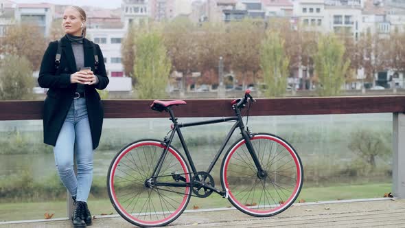 Woman standing in city by bicycle with earphones and coffee cup