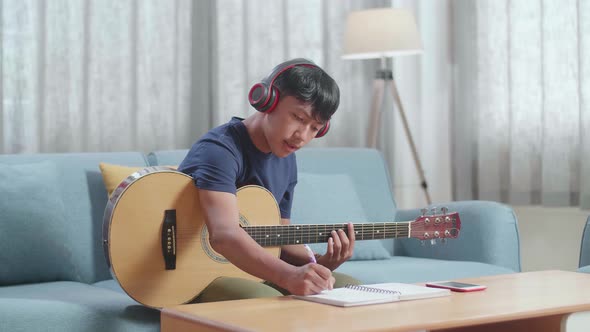 Asian Boy Composer Wearing Headphones With Guitar And Composing Music On Note Book At Home