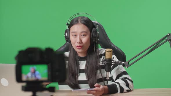 Asian Woman With Headphone Playing Game On Mobile Phone While Shooting Video On The Green Screen