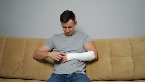 Young Man Scratches Forearm in Gypsum Band Frowning on Sofa