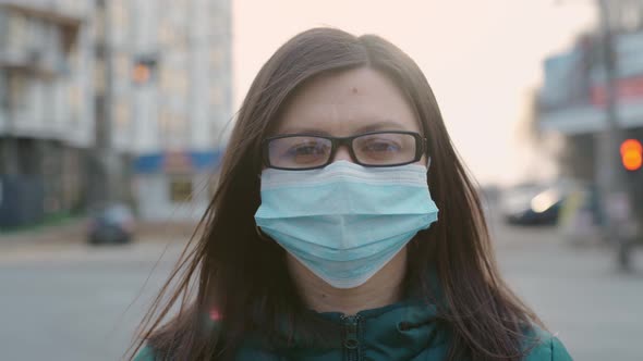 A Young Girl in Glasses and a Medical Mask Stands on the Street