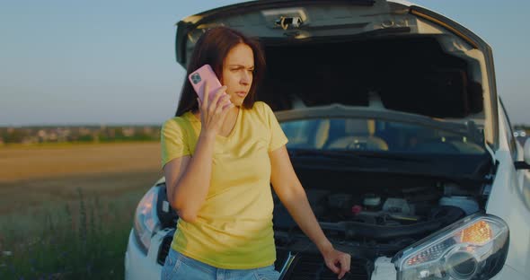 A Young Upset Angry Girl Stands By a Broken Car with an Open Hood and Talks on the Phone Waiting for