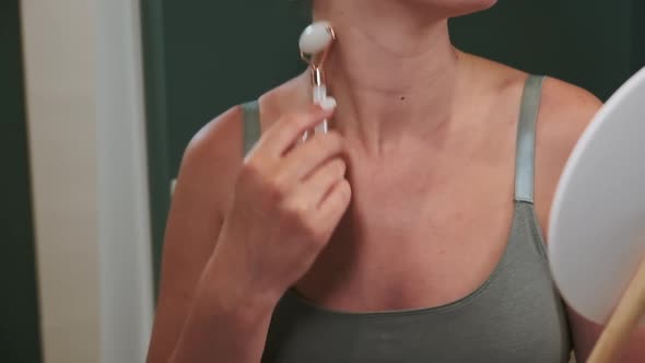 Young Woman Using Jade Facial Roller for Face and Neck Massage Sitting By the Bathroom Mirror Slow