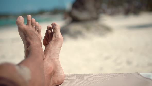 Legs Man On Beach Sun Lounger Sandy Shore Sunbathing. Tan Guy Relaxing Sandy Beach In Sunbed.