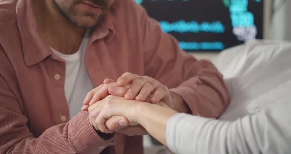 Close Up of Adult Son Holding Hands of Sick Aged Mother Lying in Hospital Bed