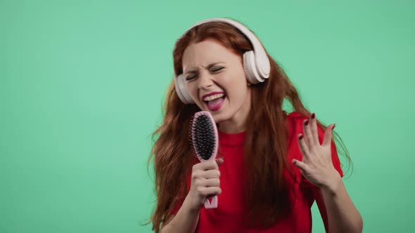 Pretty Girl with Red Hair Singing and Dancing with Brush or Comb Instead Microphone at Green Wall