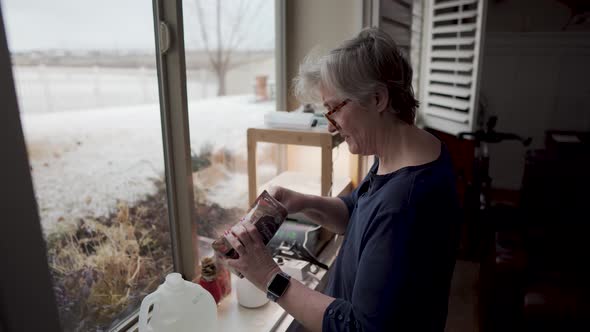 Mature woman adds fertilizer to the water used in her hydroponics indoor garden - snow outside the b