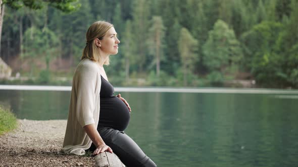 Pregnant woman touching gently her tummy when sitting near the mountain lake