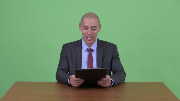 Happy Bald Multi Ethnic Businessman Showing Clipboard and Giving Thumbs Up Behind Desk