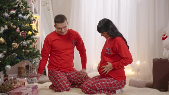 Smiling Husband Giving Christmas Present To Excited Pregnant Wife in Red Pajamas