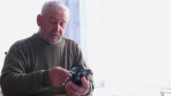 Old grandpa uses a magnifying glass to read from his smartphone.