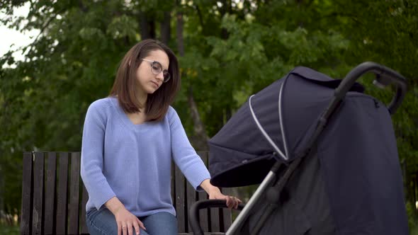 A Young Mother Sits on a Bench in the Park with a Stroller