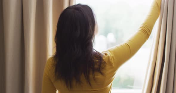 Back view of biracial woman revealing curtains and looking outside window
