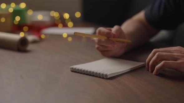 Orbit Shot of Man Writes Christmas Gift List for Holidays in Notepad with a Pencil Under Warm Light