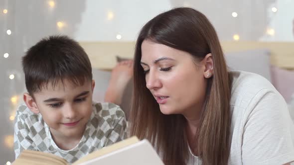 A Beautiful Girl with Long Hair Reads a Book to a Small Boy Lying on a Bed and Kisses Him