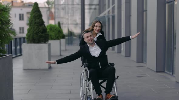 Cheerful Young Woman Pushing Wheelchair with Her Disabled Coworker While Smiling Near Office