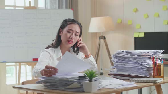 Asian Woman Talking On Phone And Taking Note While Working Hard With Documents At Office