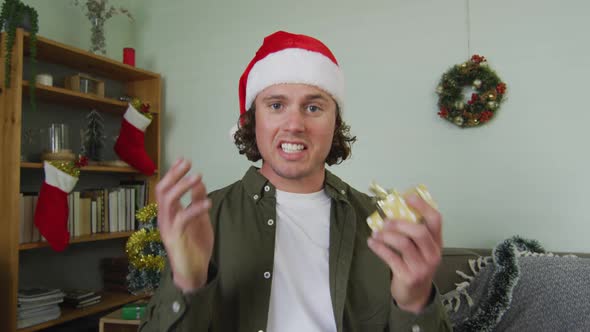 Happy caucasian man in santa hat on christmas video call holding present and talking