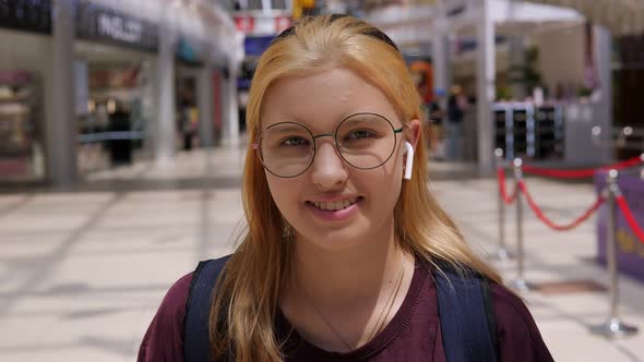 A Teenage Girl with Round Glasses and Wireless Headphones in a Shopping Center