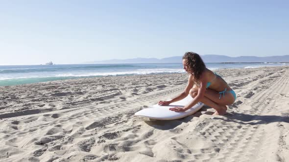 A young woman surfer waxing her board on the beach.