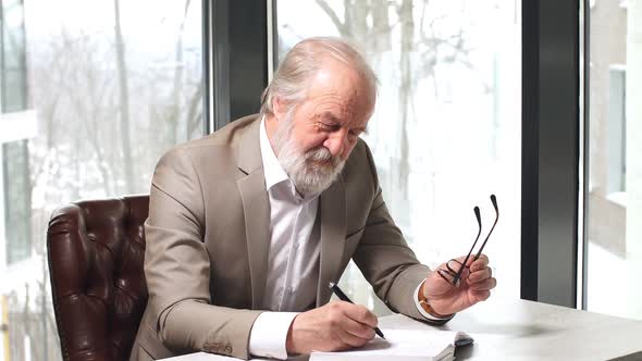 Old Professor with Grey Hair and Beard in Beautiful Suit Taking Notes in the Notebook