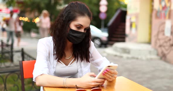 A Young Woman in a Black Mask From the Virus Sits on the Street
