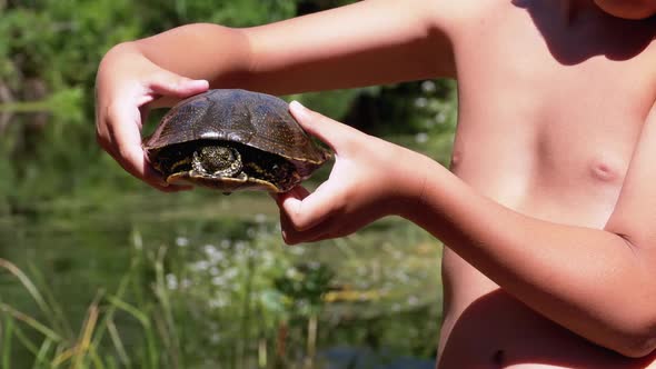 Boy Holds Turtle in Arms on Background of a River with Green Vegetation