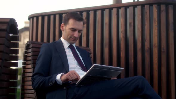 Businessman in Official Dark Blue Suit Sits on Bench in Park Before Meeting with Partners and is