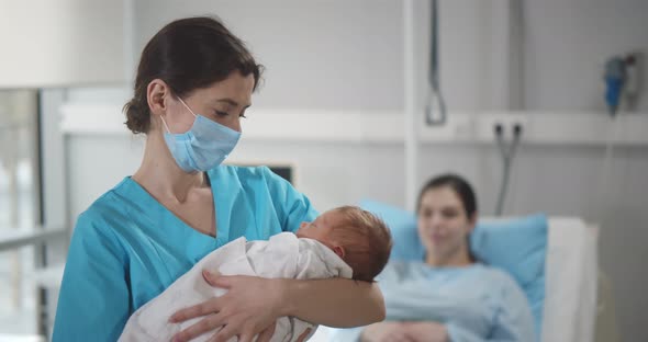 Portrait of Nurse Wearing Safety Mask Cradling Newborn in Arms with Young Mother Resting in Bed