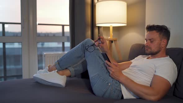 Man with Glasses Lying on the Couch in a Cozy Room and Using Smartphone for Surfing Internet or