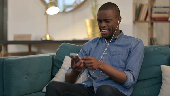 Young African Man Listening Music on Smartphone on Sofa