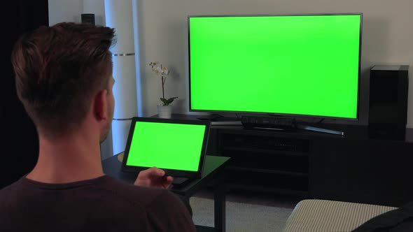 A Man, the Back of His Head Turned To the Camera, Watches a Tablet and a TV, Both with Green Screen