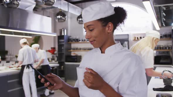 African American female chef wearing chefs whites in a restaurant kitchen using a phone and smiling