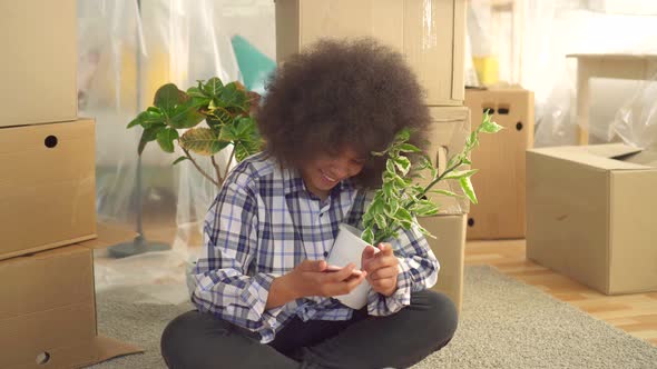 Black Woman with a Plant in His Hand Uses the Phone Sitting on the Floor Next To the Boxes To Move