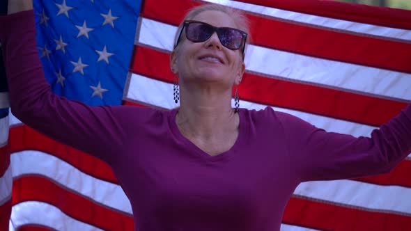 Closeup of blonde woman looking at camera and raising an American flag behind her with smiling expre