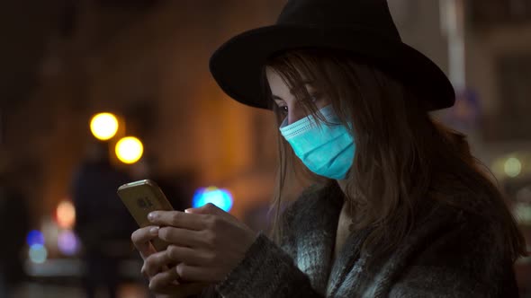 Woman in Coat and Hat Standing Using Smartphone on City Street with Bokeh Lights Shining at Night