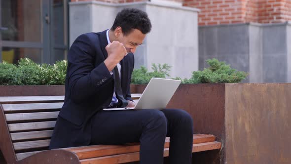 Excited African Businessman Celebrating Success on Laptop Sitting on Bench